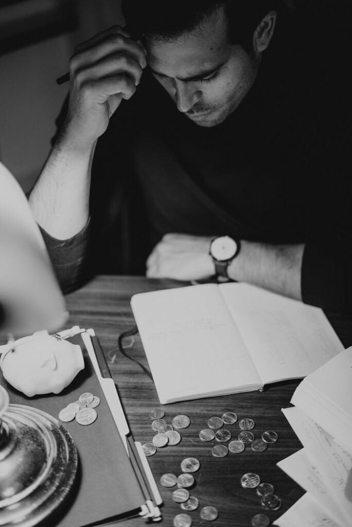 Black and white image of a man contemplating finances at a desk with coins and notebook.